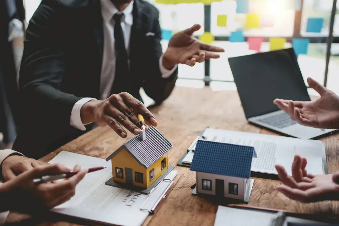 An agent across a desk from two clients, model houses and a contract between them